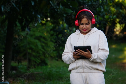 A woman (mother) is looking at a tablet while standing alone in the park, wearing red headphones.
