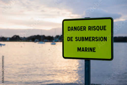 Bright yellow French coastal flooding warning sign reading “danger risque de submersion marine”, with blurred harbor and boats in soft evening light in background.