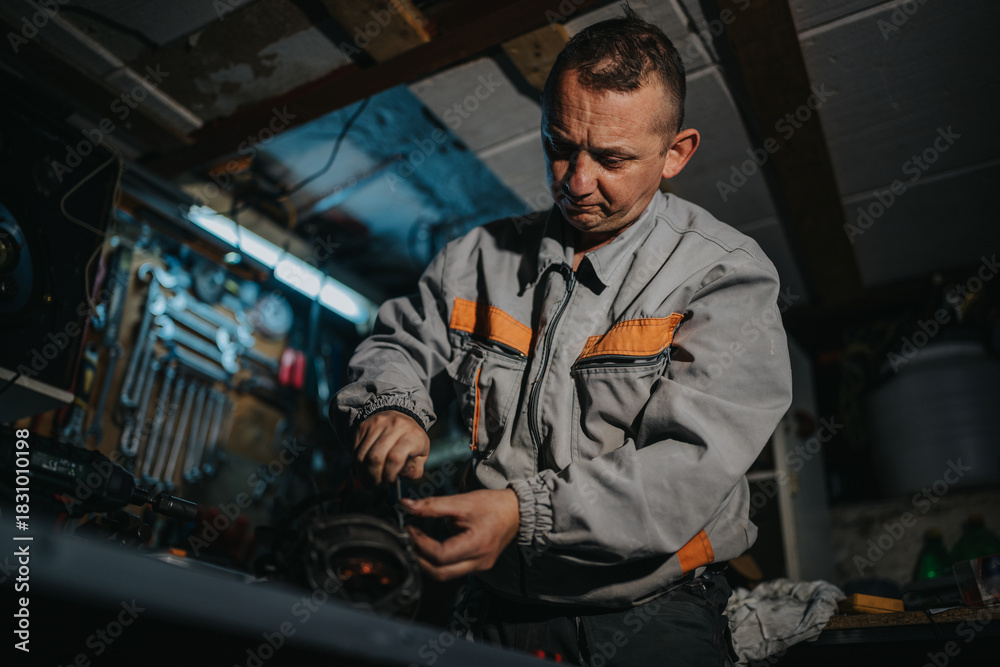 Naklejka premium A mechanic in a gray jumpsuit with orange accents concentrates on repairing a mechanical part in a busy workshop filled with tools, cables, and equipment, conveying skilled handiwork.