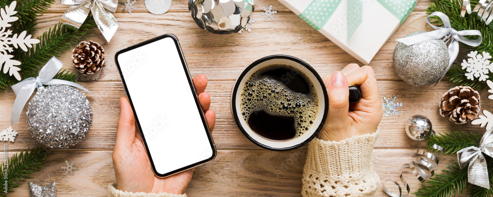 Obraz premium Above view of female hand holding smart phone with hot cup of coffee. Christmas decoration on colored background