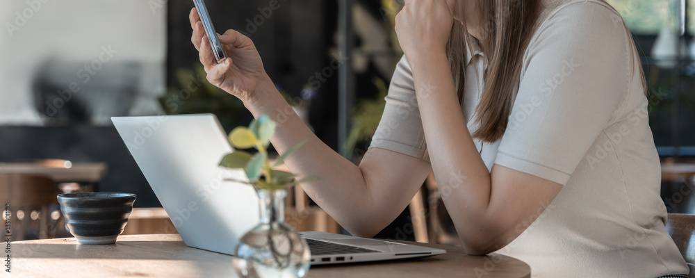 Obraz premium Woman Using Smartphone While Working On Laptop At Table In Bright Café Interior