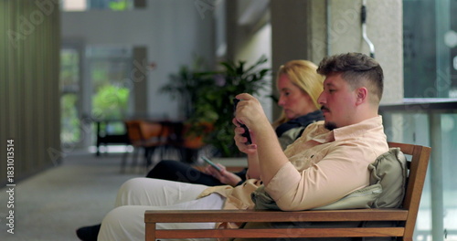 Slika na platnu Man and woman sitting apart in modern indoor hallway using smartphones, both imm