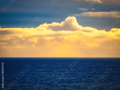 A vast, calm ocean stretches to the horizon. Golden sunlight shines through the clouds, creating a warm glow over the dark blue water during sunset.