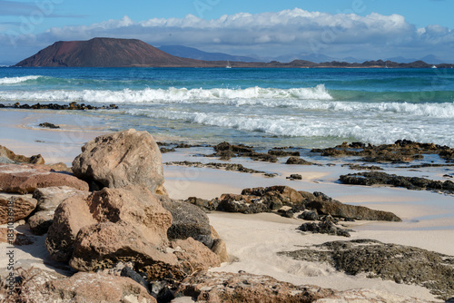 Lobos Island seen from the beach at Corralejo, Fuerteventura (Canary Islands). There are rocks and sand in the foreground. Mountains on the island of Lanzarote can be seen in the background.