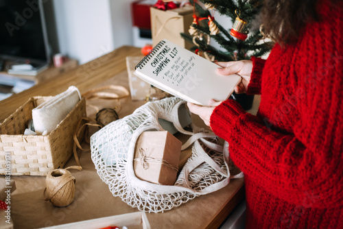 Young adult woman in red sweater reads handwritten Christmas shopping list on notebook next to small decorated tree. Christmas checklist, organized celebration, planning ahead, mindful gifting.