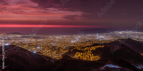 Panoramic night view of Lima city lights and coastline seen from the arid hills of the outskirts during a dramatic red sunset, highlighting urban sprawl and population density.