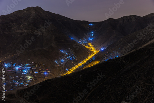 Night aerial view of a winding illuminated road cutting through dark arid mountains, highlighting the expansion of public lighting in precarious informal settlements in Lima.