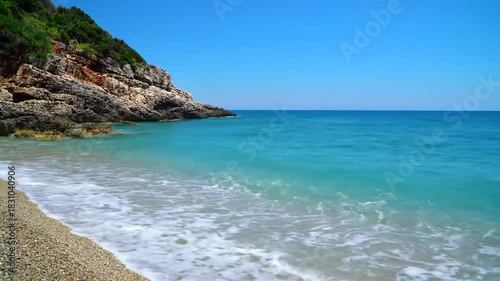 Ocean wave on pristine beach with rocky cliff and clear blue sky