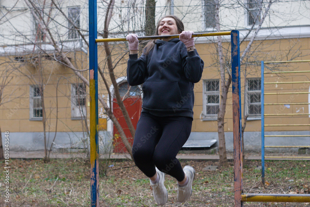 Obraz premium In an urban park, a woman lifts herself on a pull-up bar, knees bent and feet off the ground. Her face is focused, showing effort as she performs the exercise