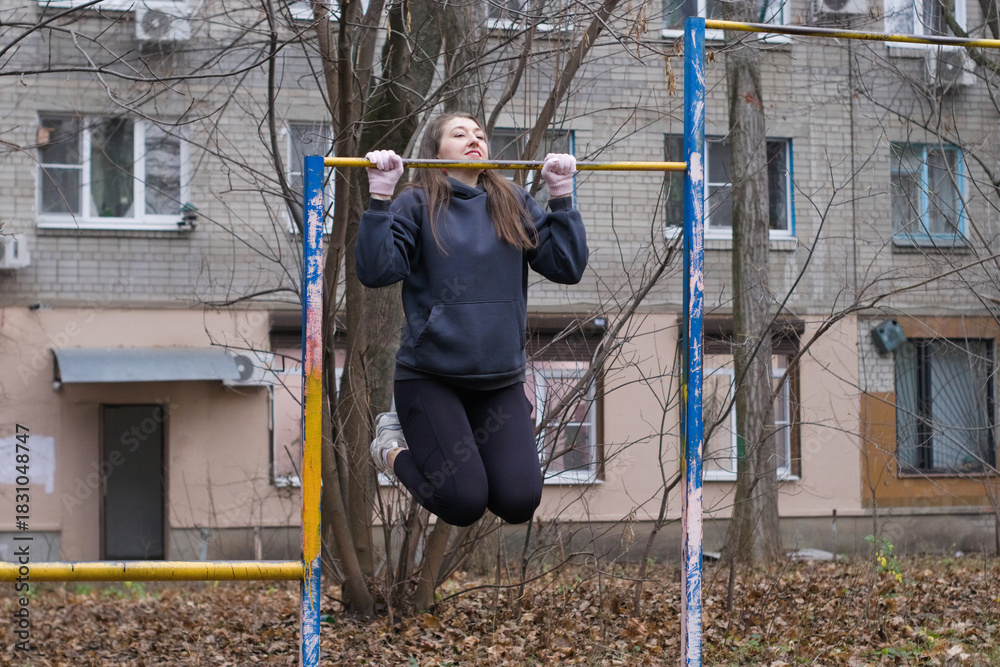 Naklejka premium European woman holds chin up position on pull up bar in city park, focusing on upper body and arm strength. Outdoor calisthenics workout, autumn setting, physical training and self improvement.