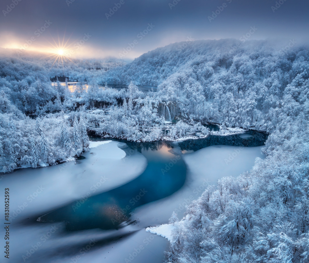 Naklejka premium Aerial drone view of frozen lakes and waterfalls surrounded by snowy forest at sunrise in Plitvice Lakes National Park, Croatia. Winter landscape with icy cascades and scenic European nature. Top view