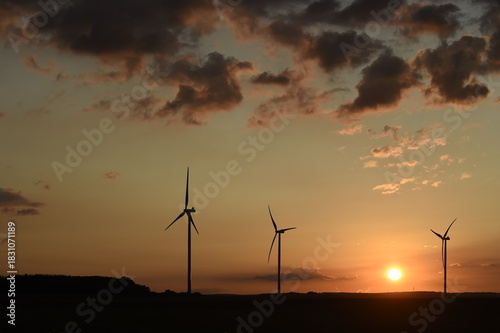 lever de soleil sur des éoliennes à contre-jour; ciel dégradé couleur bleue et orange avec des petits nuages cumulus blancs gris et noirs