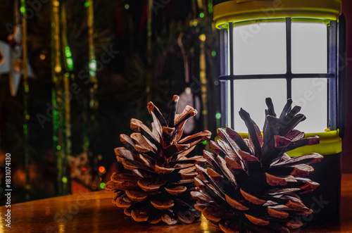Camping lantern illuminating pine cones near Christmas tree lights
