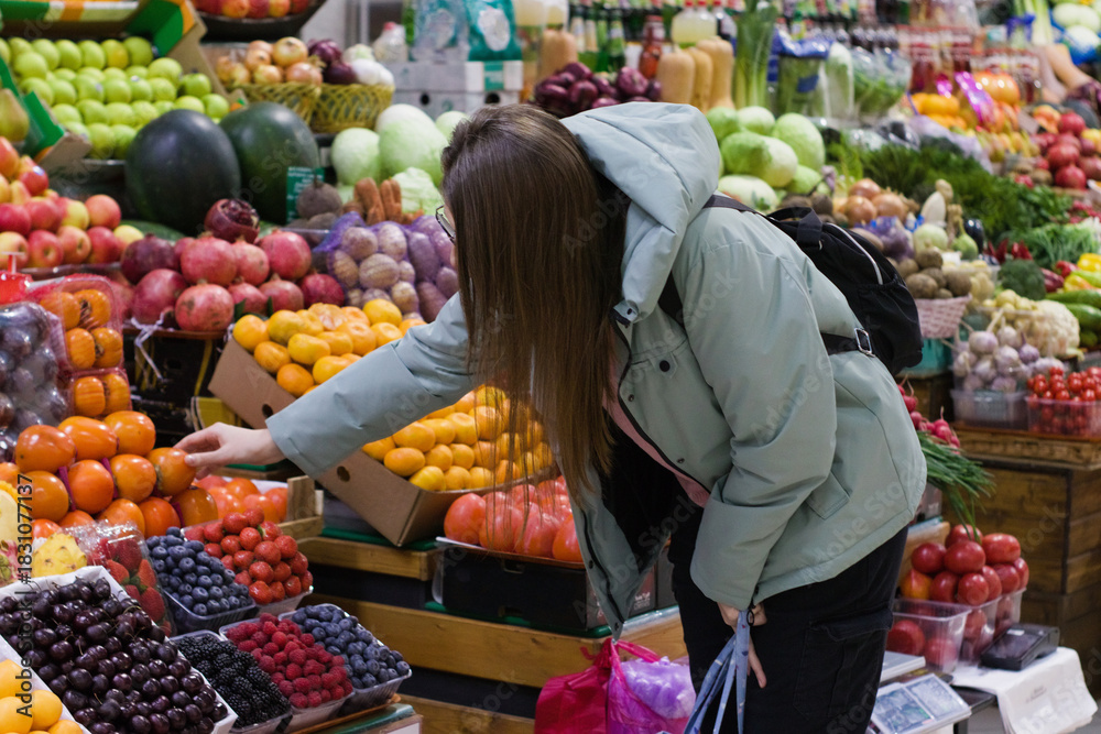 Obraz premium At a busy produce market, a young woman in a light green jacket reaches out to pick up fruit from a colorful selection. The scene is lively with a variety of fruits around her.