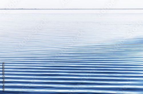 Canvas-taulu linear blue ripples in calm water dissipate at the shoreline with horizon