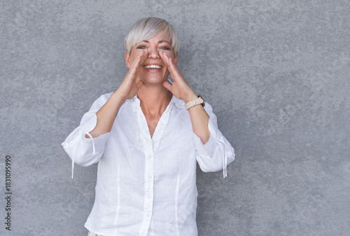 Amused woman yelling, against gray background - wall. Happy middle age woman holding hands near his mouth