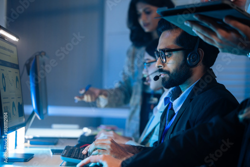 Customer service agent wears a headset talking to a client in a dark call center. Support, IT helpdesk staff, and late night corporate operations and assistance telemarketing