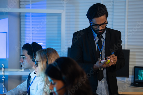 Business manager takes notes while overseeing a team of customer service agents working late in command center or call center office. high-pressure operation involving professional communication