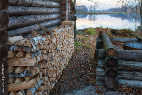 A woodpile set up against a log wall near a pond on a cloudy autumn day. Firewood stacked in a woodpile.