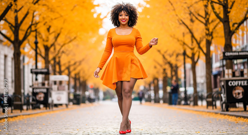 Confident woman with natural curly hair walking down a tree-lined city street in autumn, wearing a bright orange long-sleeve dress and red high heels, smiling at the camera