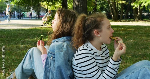 Teen girls relax in green park enjoying quick snack together sitting on grass. Teenager chews sandwich while sister watches surroundings quietly holding food