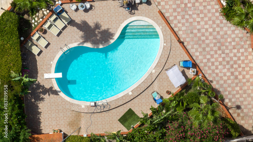 Aerial perpendicular view of a circular swimming pool on a beautiful sunny summer day. Sun loungers and deck chairs surround the pool. The pool and surrounding garden are empty.