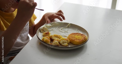 Child eats cheesecakes and twirls fork in hand spending time in kitchen. Preschooler chews food and aims cutlery at cheese pancake on frying pan surface
