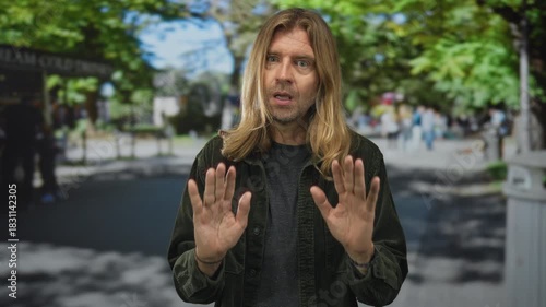 Blond man with long hair wearing green jacket holds both palms forward on park walkway under leafy trees; caution.