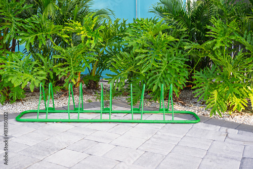 A green bike rack stands ready on smooth stone pavers, framed by a lush wall of vibrant green vegetation in the background.