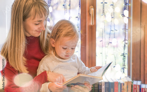 Mom and daughter are reading a book by a window decorated with garlands