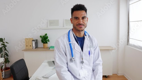 Portrait of confident young adult Latin American male doctor looking at camera standing at his medical consultation in hospital. Medical professionalism and positivity concept.