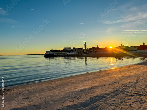 Fototapeta Naklejka Na Ścianę i Meble -  A tranquil sandy beach meets the calm water of the sea during a vibrant golden sunset. The scenic view features the silhouetted historic skyline and bell towers of Rab town against a clear blue sky.