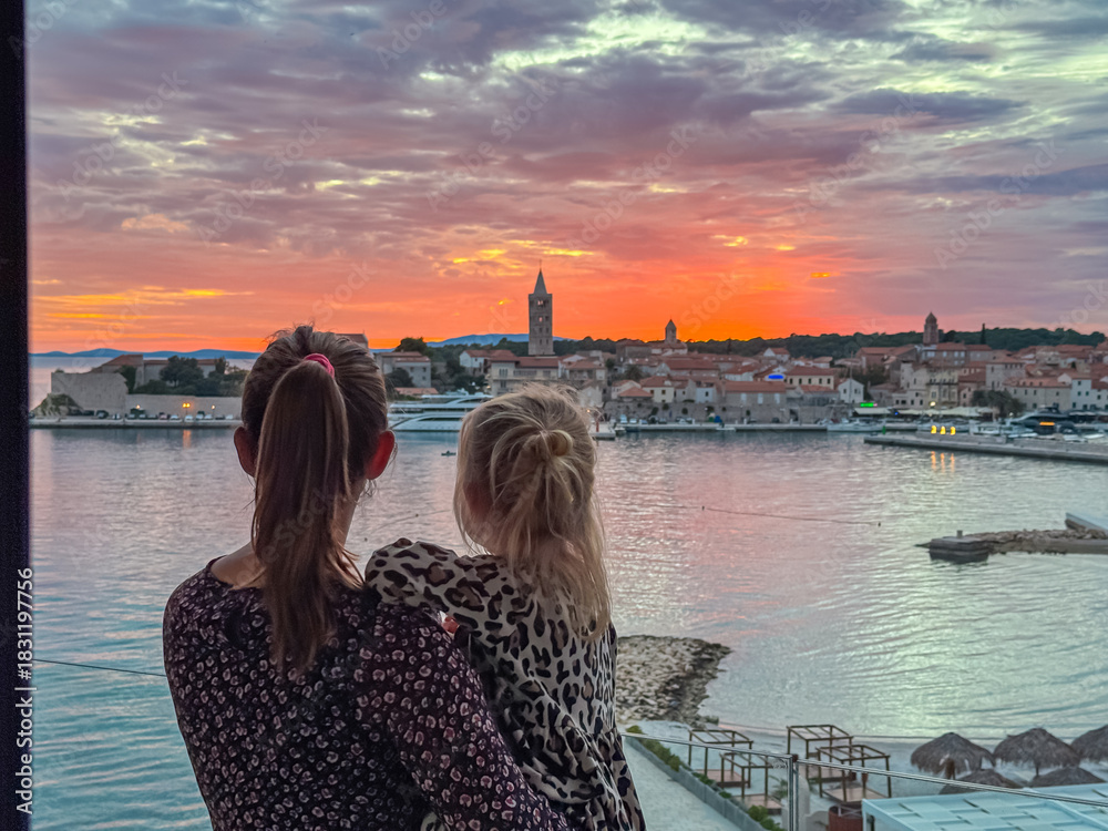 Naklejka premium A mother holds her young daughter while standing on a balcony to watch the vibrant red sunset over the historic skyline of Rab Old Town during a summer family vacation in coastal region of Croatia.