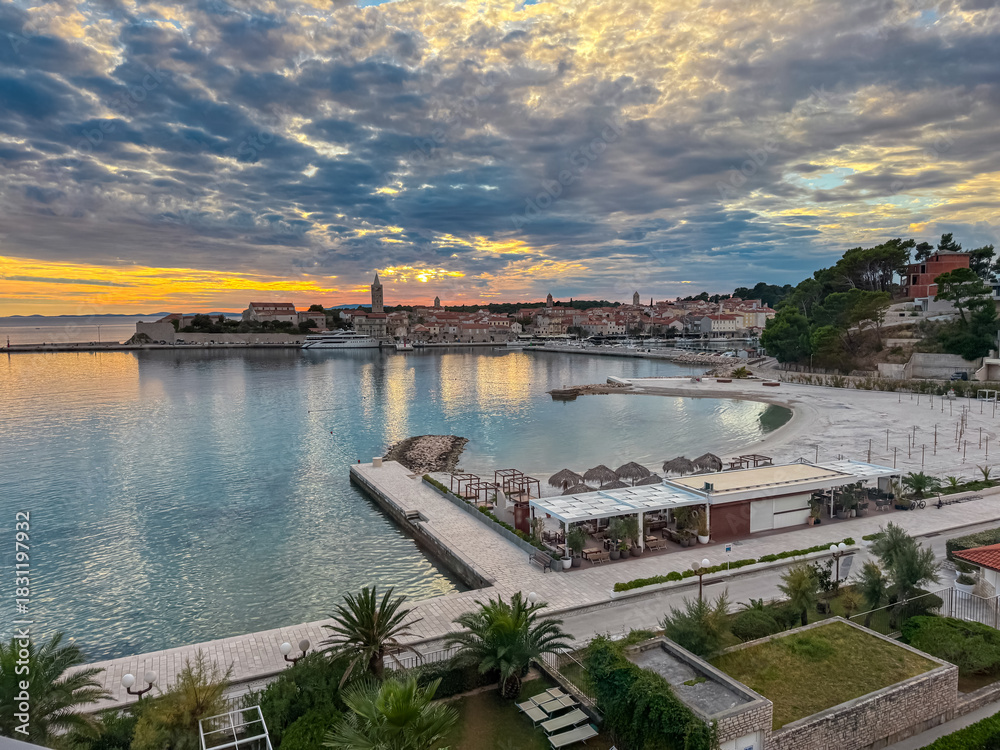 Obraz premium A scenic aerial view captures the calm turquoise bay and modern beach resort terrace facing the historic bell towers of Rab Old Town during a colorful sunset in the Kvarner region of coastal Croatia.