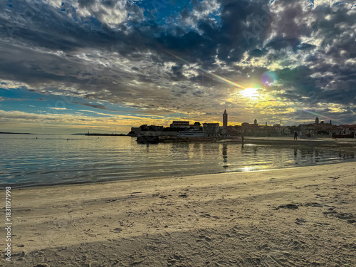 Fototapeta Naklejka Na Ścianę i Meble -  The golden sun bursts through dramatic clouds above the historic skyline of Rab Old Town while reflecting on the calm sea and sandy beach during a scenic summer sunset in coastal region of Croatia.