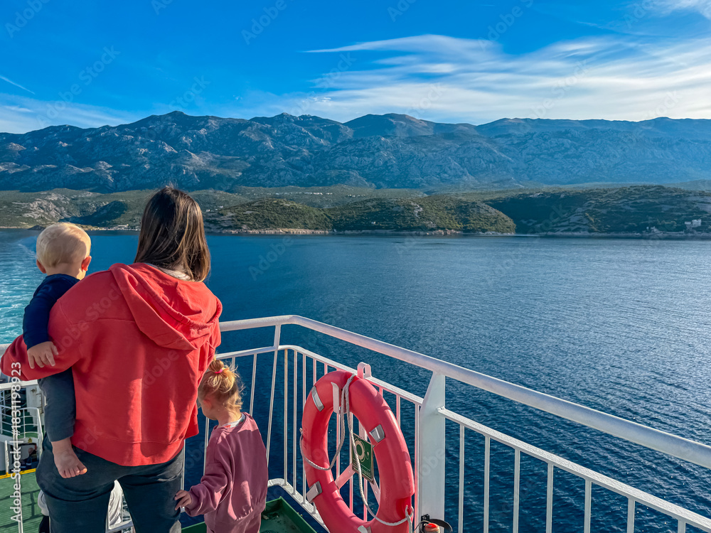Fototapeta premium A mother holding her toddler and standing with her daughter admire the majestic Velebit mountains from a ferry deck crossing blue Adriatic Sea towards Rab Island, Croatia, on a sunny family vacation.