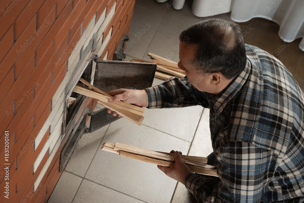 Naklejka premium Placing thin kindling into brick stove by caucasian adult man in checkered shirt while preparing fire inside warm indoor room. Winter Firewood Routine Preparing and Lighting Wood Stove