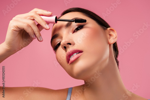 Stunning woman applying mascara on her eyelashes with a brush on a pink background, showcasing beauty and self-care