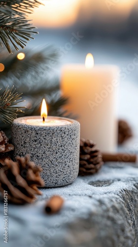 Close-up of lit candles, pine cones, and evergreen branches in a snowy winter setting. The warm glow of the candles creates a cozy and festive atmosphere.