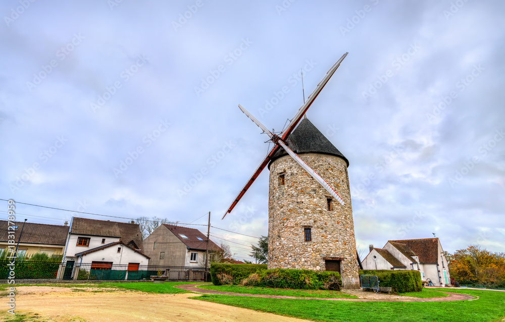 Fototapeta premium The historic Moulin de Montfermeil or Sempin Windmill in France. The stone structure with wooden blades stands in a park, surrounded by houses under a cloudy sky