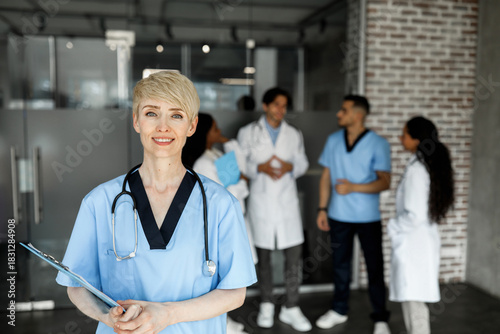 Attractive short-haired blonde woman in workwear doctor attending medical conference, smiling at camera, international group of doctors chatting while training, exchanging experience