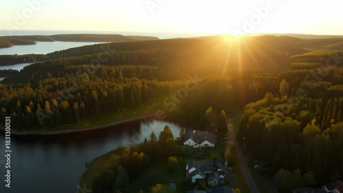 Golden sunlight bathes the lush green forest and tranquil lake, creating a serene atmosphere at dusk. The landscape unfolds with vibrant colors, inviting peaceful reflection and jo