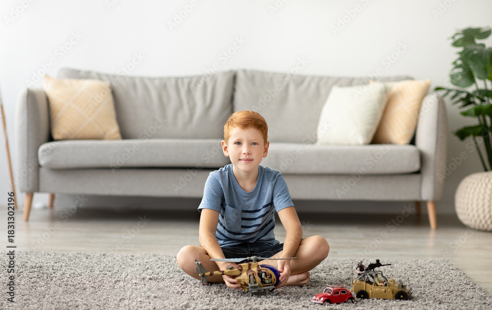 Naklejka premium A cheerful redhead boy sits comfortably on a soft carpet in his living room. He is playing happily with a helicopter and toy cars, fully enjoying his new toys and the cozy atmosphere of home.