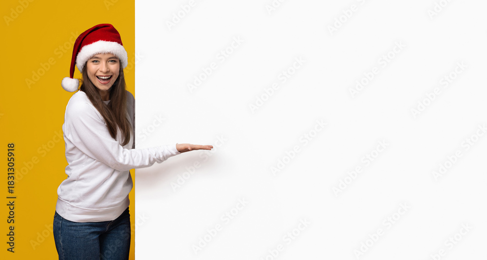 Fototapeta premium A young woman wearing a Santa hat is smiling and gesturing toward a blank space on a colorful background. She appears enthusiastic about a holiday event or promotion, embodying festive spirit.