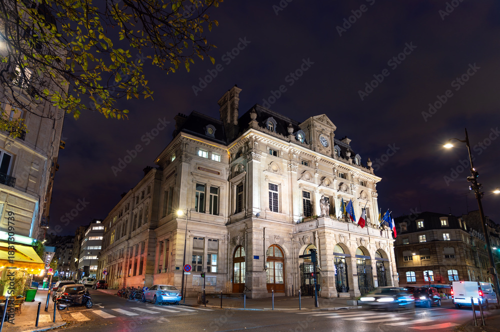 Naklejka premium Town Hall of the 18th arrondissement in Paris, France. The historic building with a clock tower and flags stands on Place Jules Joffrin at night