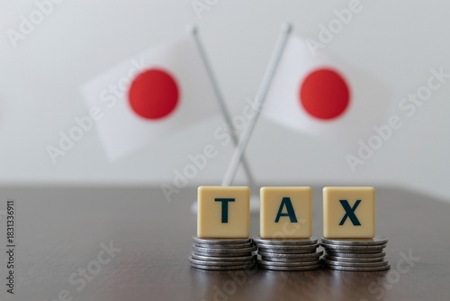 Stacks of coins topped with TAX letter blocks in focus, with two Japanese flags blurred in the background, symbolizing Japan finance, taxation, economy, investment, savings, and fiscal policy themes.