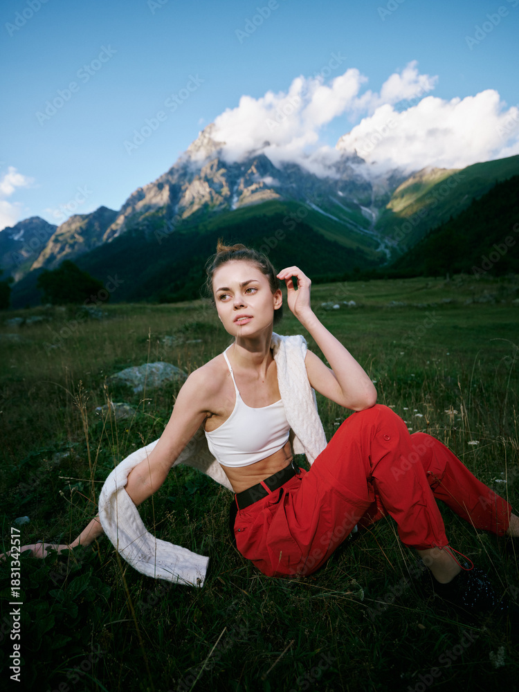 Naklejka premium Meadow scene with mountain backdrop, a woman sits on grass in a white top and red pants, a towel draped over her shoulder, serene outdoor mood under a blue sky