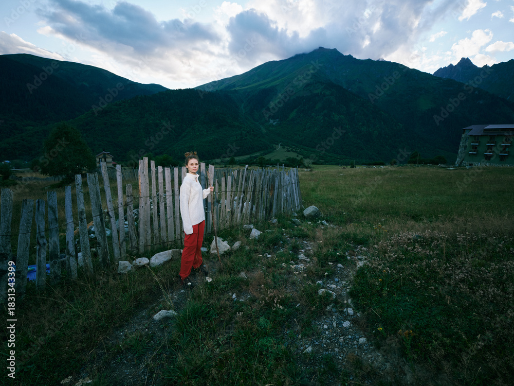 Fototapeta premium Child standing near a rustic wooden fence in a wide meadow, mountains looming in the background, moody clouds above, peaceful countryside scene with open skies.