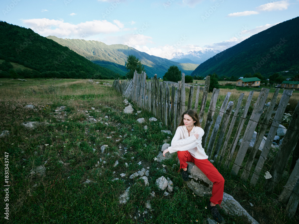 Fototapeta premium Landscape of rugged countryside with distant mountains, a wooden fence along a rocky meadow, a woman crouching beside stones, white top and red pants, clear sky and tranquil outdoor mood