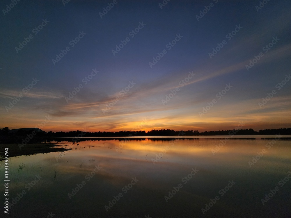 Fototapeta premium Winter sunset over a calm reservoir with golden reflections and soft clouds. Peaceful evening sky glowing with warm light across the still water.